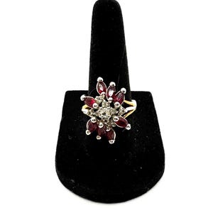 May include: A gold-toned ring with a floral design. The ring features a cluster of dark red marquise-cut gemstones surrounding a central cluster of smaller, clear gemstones. The ring is displayed on a black ring holder.