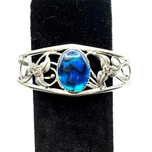 May include: Silver-toned cuff bracelet with an oval, iridescent blue gemstone center. The bracelet features openwork floral and leaf designs on the sides. The gemstone has shades of blue and green.