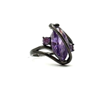 May include: A dark gray ring featuring a large, marquise-cut purple gemstone. Two smaller purple gemstones are set on the ring's band. The ring has a unique, swirling design.