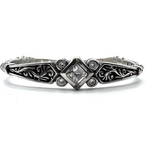 May include: A silver-toned bracelet with an ornate design. The center features a square, clear gemstone, flanked by smaller round gemstones. Intricate scrollwork adorns the bracelet's sides, creating a vintage or antique aesthetic. The bracelet is set against a white background.