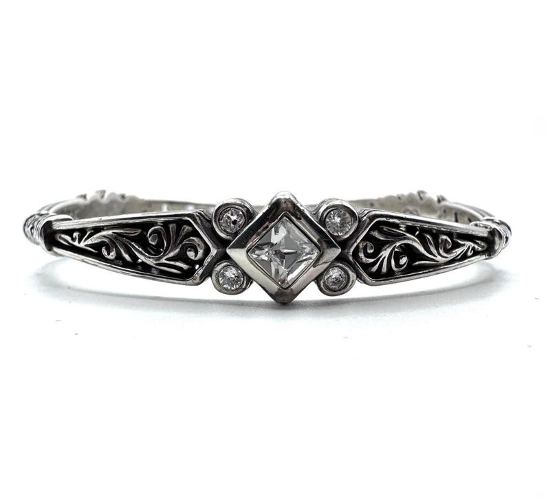 May include: A silver-toned bracelet with an ornate design. The center features a square, clear gemstone, flanked by smaller round gemstones. Intricate scrollwork adorns the bracelet's sides, creating a vintage or antique aesthetic. The bracelet is set against a white background.
