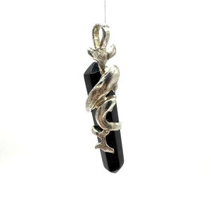 May include: A silver-toned pendant featuring a polished, black, rectangular gemstone. The gemstone is wrapped with a decorative silver metal design. The pendant hangs from a thin, silver-colored string against a white background.