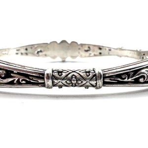 May include: A silver bangle bracelet with intricate filigree and floral designs. The bracelet features a central decorative element with a repeating pattern. The metal has a polished finish, reflecting light and highlighting the detailed craftsmanship.
