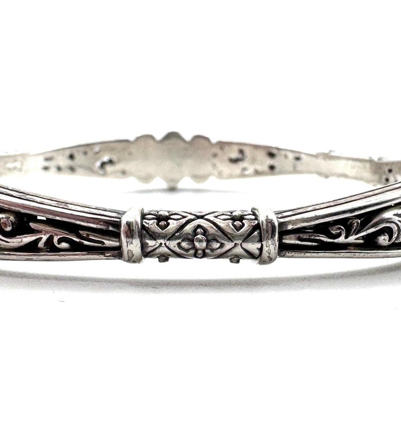 May include: A silver bangle bracelet with intricate filigree and floral designs. The bracelet features a central decorative element with a repeating pattern. The metal has a polished finish, reflecting light and highlighting the detailed craftsmanship.