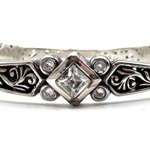 May include: A silver-toned bracelet with an ornate design. The bracelet features a central square-cut clear gemstone, surrounded by smaller round clear gemstones. Intricate scrollwork and decorative elements adorn the band, creating a vintage aesthetic.