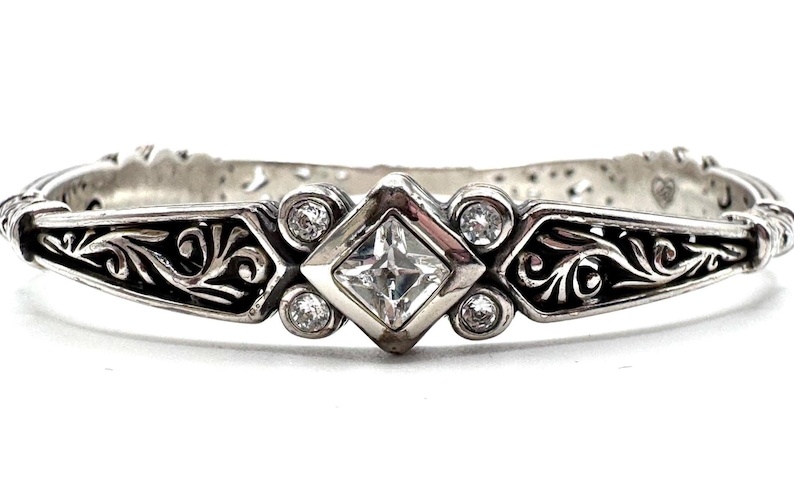 May include: A silver-toned bracelet with an ornate design. The bracelet features a central square-cut clear gemstone, surrounded by smaller round clear gemstones. Intricate scrollwork and decorative elements adorn the band, creating a vintage aesthetic.