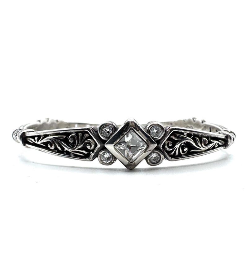 May include: A silver bracelet with an ornate design. The bracelet features a central square-cut clear stone, surrounded by smaller round stones. The band has a detailed, swirling pattern. A classic piece of jewelry.