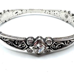 May include: Silver-toned bracelet with a central square-cut clear gemstone and smaller round clear gemstones. The bracelet features ornate scrollwork and decorative detailing. The bracelet is approximately 7 inches in circumference.
