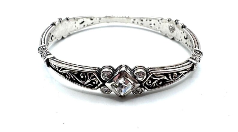 May include: Silver-toned bracelet with a central square-cut clear gemstone and smaller round clear gemstones. The bracelet features ornate scrollwork and decorative detailing. The bracelet is approximately 7 inches in circumference.