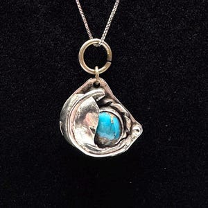 May include: A silver pendant necklace featuring a turquoise stone set within a stylized, openwork design. The pendant hangs from a delicate silver chain. The pendant's design has a unique, organic shape.