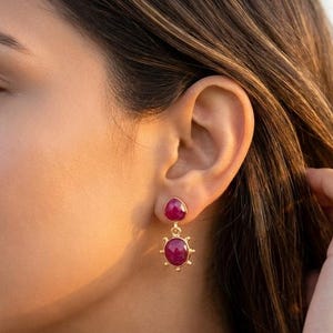 May include: A pair of gold drop earrings featuring two round, deep pink cabochon stones. The top stone is smaller and sits directly on the earlobe, while the larger bottom stone is suspended by a small gold link and framed by tiny gold spikes.