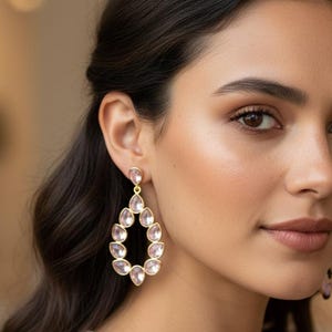 May include: Gold-toned teardrop earrings with a cluster of light pink stones. The earrings are worn by a person with dark hair and a neutral-toned top. The earrings have a delicate, elegant design.