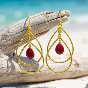 May include: Gold-tone teardrop earrings with a textured finish. Each earring features a double teardrop design with a small, oval, deep red gemstone suspended in the center. The earrings are displayed on driftwood against a blurred beach background.