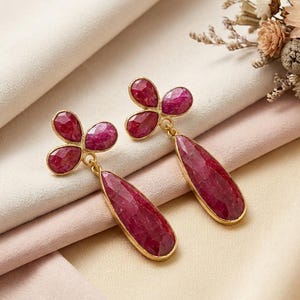 May include: A pair of drop earrings featuring faceted, deep red ruby-colored stones set in gold-toned metal. Each earring consists of a three-petaled floral-shaped top connected to a long, teardrop-shaped pendant that hangs vertically from the base.