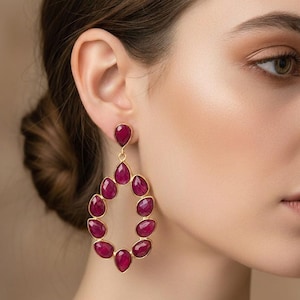 May include: A close-up of a woman wearing a pair of gold-colored teardrop earrings. Each earring features a series of vibrant, oval-shaped, ruby-red gemstones. The earrings are a striking accessory, adding a pop of color and elegance.