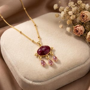 May include: A gold chain necklace featuring a central oval-shaped faceted deep red gemstone pendant. Three smaller, light pink, pear-shaped faceted gemstones hang from the bottom of the main oval stone, creating a delicate, dangling design.