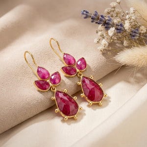 May include: A pair of gold drop earrings featuring faceted, deep pink, pear-shaped and oval-shaped stones. Each earring has a hook-style closure and a cluster of stones arranged in a vertical design with small gold prongs securing each gemstone.
