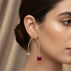 May include: Gold-toned earrings with a textured finish. One earring features a triangular white gem, while the other has a square, deep red gemstone. The earrings have an arched design, creating a modern and elegant look.