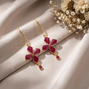 May include: A pair of gold drop earrings featuring a cross-shaped design composed of four deep magenta, faceted stones set in gold frames, with a single small teardrop-shaped magenta stone dangling from the bottom of each earring.