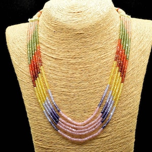 Peut inclure: Collier de perles à plusieurs rangs présenté sur un mannequin. Le collier présente des rangées de perles colorées dans les tons de rose, violet, jaune, vert et rouge. Les perles sont petites et espacées uniformément, créant un aspect texturé. Le fond est noir.