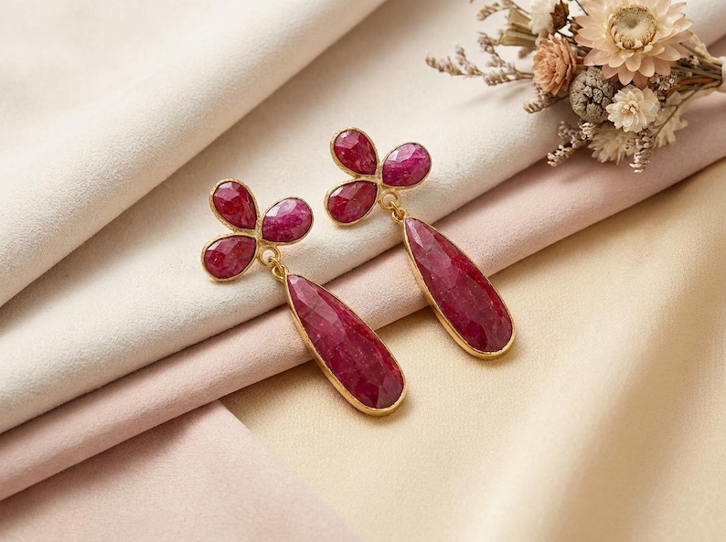May include: A pair of drop earrings featuring faceted, deep red ruby-colored stones set in gold-toned metal. Each earring consists of a three-petaled floral-shaped top connected to a long, teardrop-shaped pendant that hangs vertically from the base.