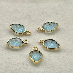 May include: Five leaf-shaped pendants with a light blue gemstone and gold-colored trim. Each pendant has a small loop at the top for attaching to a chain or other jewelry component. The gemstones have a textured surface.