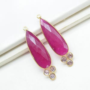 May include: Two elongated, faceted, magenta-colored gemstone pendants with gold-colored trim. Each pendant has a small loop at the top and a cluster of three smaller, light pink gemstones at the bottom. The pendants are displayed on a white surface.