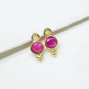 May include: Two gold-coloured jewellery charms, each featuring a round, faceted, magenta-coloured stone. Each charm has a small loop at the top and a cluster of three small beads at the bottom, set against a white background.