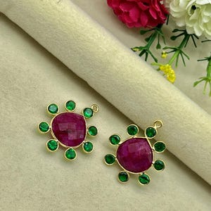 May include: Two gold-toned pendants, each featuring a large, faceted, deep red gemstone at the center. Each pendant is surrounded by eight smaller, round, green gemstones. The pendants are displayed on a light beige surface.