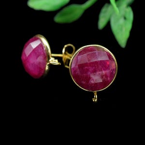 Pode incluir: Um par de brincos de ouro com grandes pedras preciosas redondas, facetadas, vermelho rubi. Os brincos são colocados sobre um fundo preto com um toque de folhagem verde no canto superior direito.