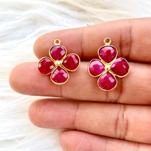 Op de afbeelding: Twee goudkleurige, vierbladige bloemvormige hangers met gefacetteerde, dieprode edelstenen. Elk bloemblad heeft een afgeronde vorm en de edelstenen zijn gezet in een goudkleurig metaal. De hangers hebben kleine lussen aan de bovenkant om op te hangen.