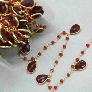 Könnte beinhalten: Nahaufnahme von Schmuck mit einer Goldkette mit kleinen roten Perlen und größeren, tropfenförmigen, dunkelroten Edelsteinen. Die Edelsteine sind in goldenen Fassungen gefasst. Der Schmuck wird auf einer hellgrauen Oberfläche präsentiert.