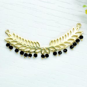 May include: A gold-colored, leaf-shaped jewelry component with a curved design. The component features a series of cut-out leaf shapes and is adorned with small, dangling black beads. The piece is designed for jewelry making.