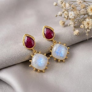 May include: A pair of gold-toned earrings featuring a faceted, pear-shaped deep red gemstone at the top, connected to a square, cabochon-cut iridescent moonstone at the bottom, which is framed by small gold beads along its edges.