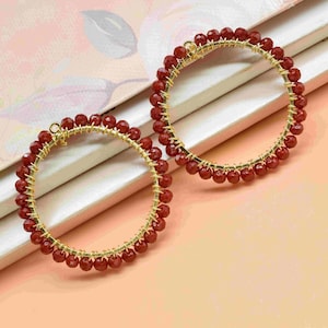 May include: A pair of gold-toned hoop earrings. Each earring is adorned with small, round, red beads. The beads are strung along the hoop's circumference, creating a textured, circular design. The earrings rest on a light-colored surface.