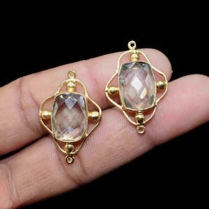 Pode incluir: Dois pingentes de pedras preciosas douradas com uma pedra central clara e facetada. Os pingentes têm um design ornamentado e vazado com pequenos detalhes dourados. As pedras preciosas são mantidas no lugar por uma moldura dourada decorativa.