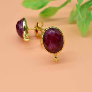 Puede incluir: Un par de pendientes de botón dorados con grandes piedras preciosas ovaladas, facetadas, de color rojo intenso. Los pendientes están colocados sobre un fondo melocotón suave con follaje verde en la esquina superior derecha.