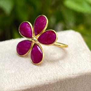 以下が含まれることがあります： 金色の指輪、花のモチーフ。 指輪には、それぞれ金色のフレームにセットされた、マゼンタ色の5つの花びらがあります。 指輪は、ぼやけた緑色の背景に対して、白い表面に表示されています。