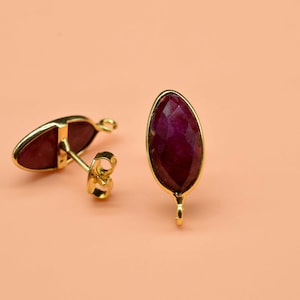 May include: A pair of gold-coloured stud earrings featuring a teardrop-shaped, deep red gemstone. The earrings are set against a peach-coloured background, with one earring lying on its side.