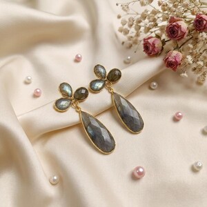 May include: A pair of gold-toned drop earrings featuring faceted, teardrop-shaped grey labradorite stones suspended from a cluster of three smaller, round labradorite cabochons. The stones exhibit iridescent flashes of blue and green.