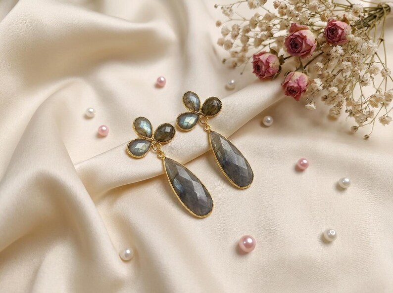 May include: A pair of gold-toned drop earrings featuring faceted, teardrop-shaped grey labradorite stones suspended from a cluster of three smaller, round labradorite cabochons. The stones exhibit iridescent flashes of blue and green.
