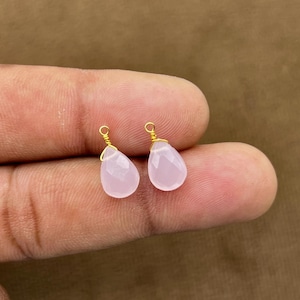 May include: Two light pink teardrop-shaped gemstone pendants with gold-colored wire wrapping. The gemstones are faceted and have a slight translucence. The pendants are displayed against a neutral background.