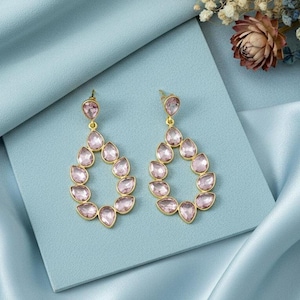 May include: A pair of gold-toned teardrop earrings with pink gemstones. Each earring features a small pink gem at the top, with a teardrop-shaped design of pink gems below. The earrings are displayed on a light blue surface with floral accents.