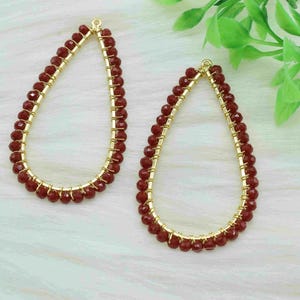 May include: Two teardrop-shaped earrings. Each earring has a gold-coloured wire frame, with a border of small, dark red beads. The earrings are displayed on a white, textured surface, with green foliage in the background.