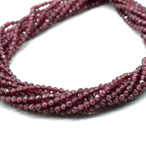 Könnte beinhalten: Stränge aus kleinen, facettierten, tiefroten Perlen. Die Perlen sind aufgefädelt und bilden mehrere leicht verdrehte Stränge. Die Perlen haben eine glänzende, reflektierende Oberfläche. Der Hintergrund ist weiß.