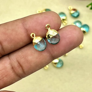 May include: Two gold-plated teardrop-shaped pendants with light blue, faceted glass stones. The pendants are held between fingers against a light yellow background. Additional pendants are scattered in the background.
