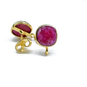 Pode incluir: Um par de brincos de botão dourados com pedras preciosas vermelho escuro de corte quadrado. Os brincos são cravejados em uma moldura dourada e possuem fecho de pino e borboleta. As pedras preciosas têm uma superfície facetada.