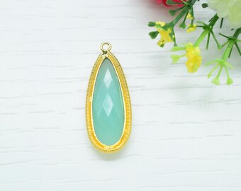 May include: A teardrop-shaped pendant with a faceted, light-green gemstone set in a gold-colored, textured frame. The pendant has a small loop at the top for hanging. The background is a white, textured surface with flowers.