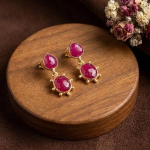 May include: A pair of gold drop earrings featuring two faceted, oval-shaped, deep pink rubies each. The stones are set in gold bezels with small decorative gold prongs around the edges, creating a sunburst effect on the lower stone.