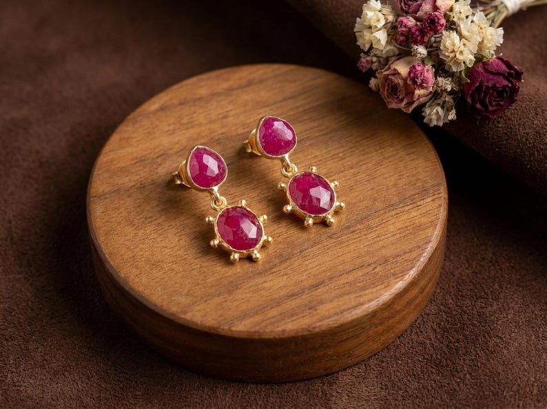 May include: A pair of gold drop earrings featuring two faceted, oval-shaped, deep pink rubies each. The stones are set in gold bezels with small decorative gold prongs around the edges, creating a sunburst effect on the lower stone.